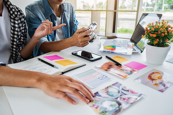 Two people discussing photography techniques while examining a camera, a notebook with notes, a smartphone, and colorful sticky notes on a table with various photographs and a potted plant in the background.