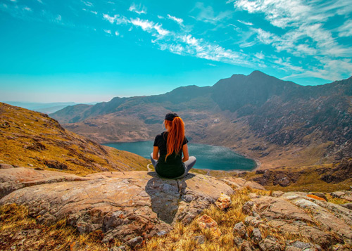 A person sitting on a rock, overlooking a serene lake surrounded by mountain ranges under a clear blue sky. Perfect for nature enthusiasts and tranquil landscapes.