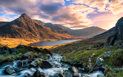 A scenic mountain landscape featuring a flowing stream in the foreground, with dramatic peaks and a serene lake nestled among golden hills under a colorful sunset sky. Beautiful natural scenery.