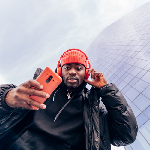 young man wearing red headphones and a red beanie, holding a smartphone, with a modern glass building in the background