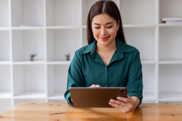 Woman in a green shirt sitting at a wooden table, looking at a tablet with a smile, in a bright room with white shelves.