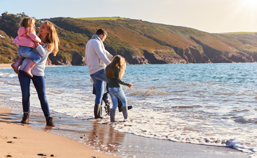 Family enjoying a sunny day at the beach, with a mother holding a young child and a father with an older child playing in the water. The scene captures a joyful moment by the ocean, highlighting family beach activities.