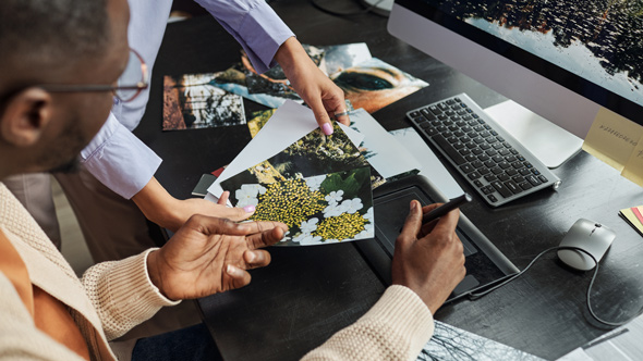 Two people collaborating at a desk, reviewing printed photographs of nature and landscapes while using a computer.