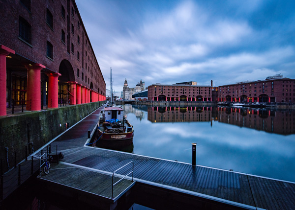 Historic docks reflecting in calm water under cloudy skies, with a boat and waterfront buildings in view.